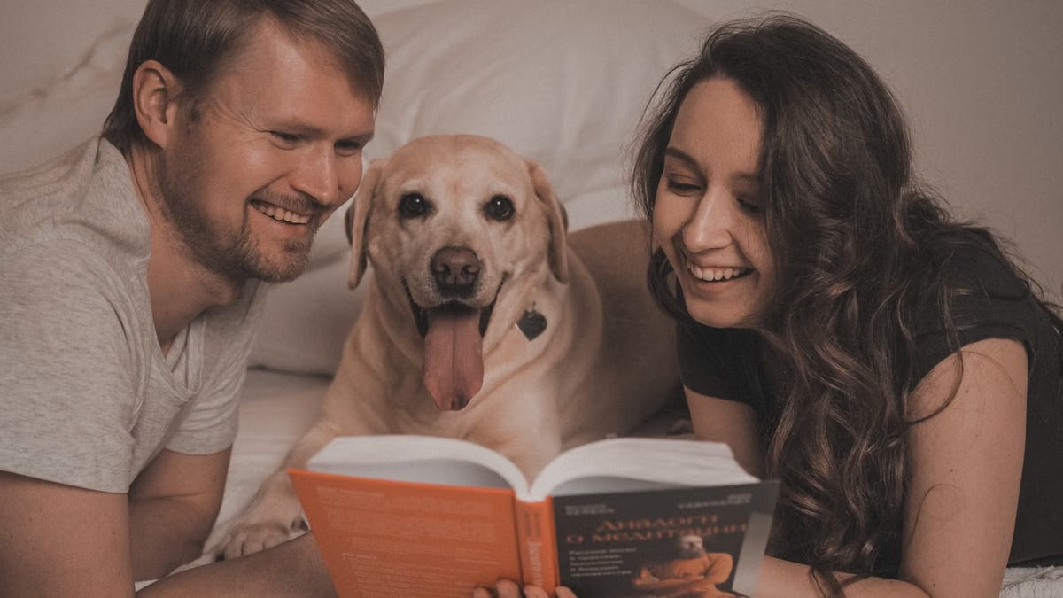 Smiling couple with their dog enjoying a book together in a cozy and relaxed bedroom setting.